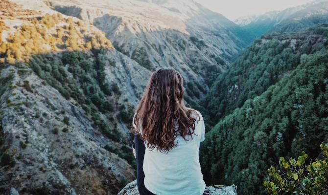 Mujer joven sentada de espaldas frente a un paisaje montañas. Photo by Helena Cook on Unsplash