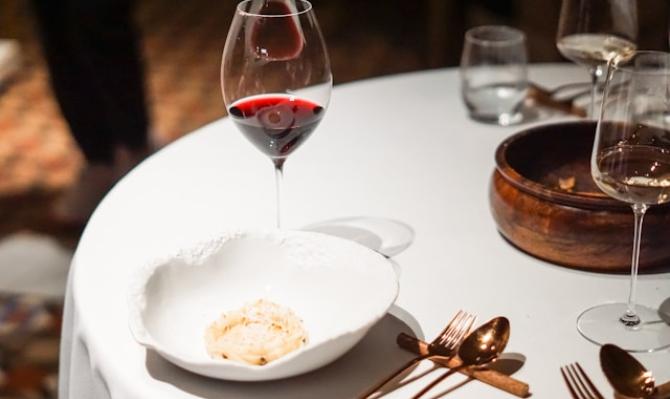 Mesa de restaurante con mantel blanco, copa de vino tinto, plato y cazuelilla de barro. Photo by Sander Traa on Unsplash