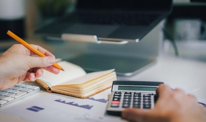 Hombre sujetando una calculadora y un bolígrafo en frente de un ordenador portátiol. Photo by Jakub Zerdzicki on Unsplash
